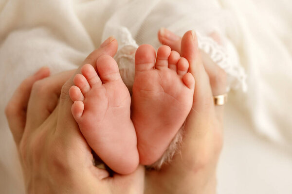 Close-up of a newborn baby tiny feet with visible toes and heels, gently held in mothers caring hands and shown to the camera on a clean milk-white background.