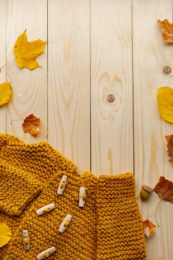 Üst görünümde, ahşap düğmeli sarı bir hırka, renkli sonbahar yaprakları ve açık ahşap arka planda meşe palamudu ile çevrili. Mevsimlik tasarım için mükemmel