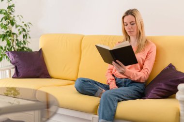 Attractive blonde girl sitting on her sofa and reading a good book, relaxing at home, enjoying leisure time clipart