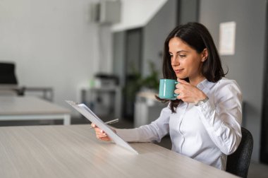 Genç ve çekici bir ofis çalışanı evrak okuyor ve bir fincan kahve içiyor.