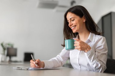 Gülümseyen genç iş kadını ofiste çay içiyor.