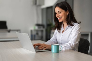 Ofiste çalışırken, masasında bir fincan kahveyle dizüstü bilgisayar kullanan gülümseyen kadın.