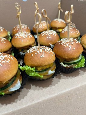 Mini burgers with fresh veggies, cheese, and sesame buns, neatly packed for parties or catering in stylish wrappers and skewers