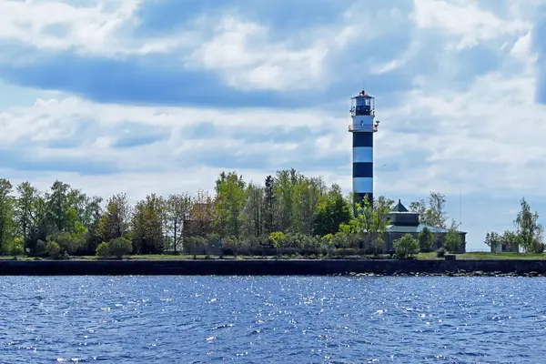 Bulutlu bir gökyüzünün altında deniz feneri. Önde gelen siyah beyaz çizgili deniz feneri, ağaçlarla çevrili, yemyeşil çimenli bir adada dimdik duruyor. Nehrin ya da denizin sakin, koyu mavi suyu ön planda uzanır, bulutlu gökyüzünden gelen ışığı yansıtır..