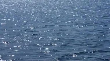 Deniz suyunun yüzeyi, sayısız güneş ışığı yansıması ile parıldıyor. Hafif dalgalanmalar dinamik ama barışçıl bir doku yaratır. Derin mavi alanda dans eden parlak ışık parıltılarıyla.. 