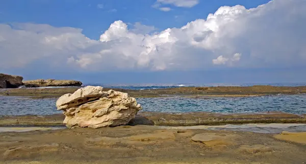 Büyük, desenli, açık renkli kaya ön plandaki düz, doğal kaya rafında dinleniyor. Bu platformun ötesinde, sakin ve berrak sular, çarpıcı beyaz kümülüs bulutlarıyla dolu, çarpıcı bir gökyüzü altında, doğal kanalları ve havuzları doldurur.. 