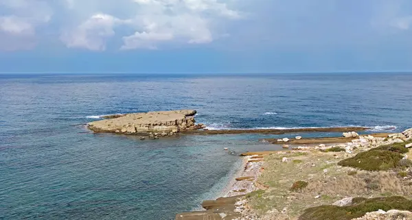 Denizin berrak, durgun mavi sularına uzanan kayalık yarımadanın yer aldığı kıyı manzarası. Ön planda küçük, gelişmemiş, seyrek bitki örtüsüne sahip kayalık bir sahil görünüyor, ince bulutlu bir gökyüzünün altındaki geniş okyanusa açılıyor..