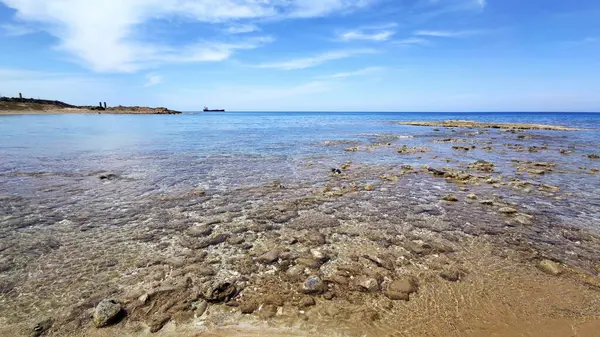 Temiz kıyı suları, şeffaf yüzeyin hemen altında kayalık ve kumlu deniz yatağı. Canlı turkuaz ve denizin mavi renkleri ufka doğru uzanıyor. Uzak bir gemi ve soluk bir kara parçası parlak, parçalı bulutlu bir gökyüzünün altında görülebiliyor.. 
