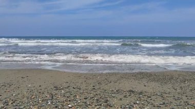 Ön planda bir çakıl taşı ve kiremit plajı barındıran kıyı manzarası denizin berrak, mavi-yeşil sularına açılır..