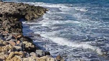 Güçlü deniz dalgaları engebeli, doğal kayalık bir kıyıya çarpıyor ve köpürüyor. Canlı mavi ve yeşil sular, denizin ham enerjisini ve dinamik gücünü gösteren beyaz köpükle çalkalanıyor.. 