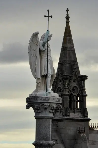 Detaylı bir sütunun üzerinde haç tutan oyulmuş taş bir melek arkasında gotik bir kule olan, bulutlu bir Dublin gökyüzüne karşı yakalanmış, inancı ve ebedi huzuru simgeleyen.