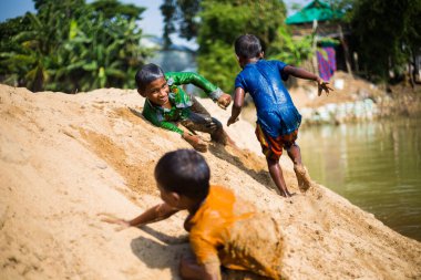 Taherpur, Bangladeş - 05 Kasım 2019: Nehir kıyısının kumlarında ıslak elbiselerle eğlenen çocuklara odaklanın. Su baskını sırasında çocuklar kum tepesinde koşup oynuyorlar..