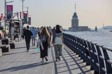 04-10-2024 İstanbul-Türkiye: Arka planda Bakire Kulesi ile rıhtım boyunca yürüyen insanlar