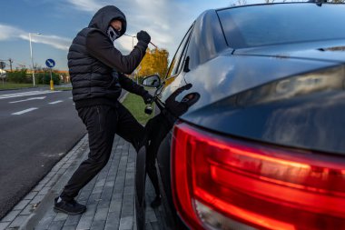 Şehir merkezinde bir bavul kullanarak araba çalmaya teşebbüs etmiş. Yüzünde maske olan kapüşonlu bir hırsız.