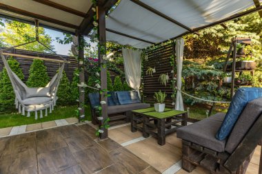 Ahşap bir bahçe çardağı, beyaz perdeli ahşap bir çardak, salıncaklı bir gevşeme alanı, terasta Brezilya hamağı.