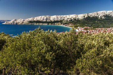 Hırvatistan 'ın Krk Adası' ndaki kayalık dağlarla çevrili turkuaz Adriyatik Denizi, marina, kumlu plaj ve kıyı kasabasının açık mavi gökyüzü altında Baska Körfezi manzaralı manzarası.
