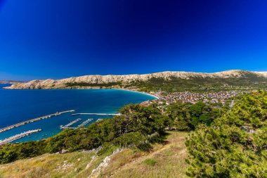 Hırvatistan 'ın Krk Adası' ndaki kayalık dağlarla çevrili turkuaz Adriyatik Denizi, marina, kumlu plaj ve kıyı kasabasının açık mavi gökyüzü altında Baska Körfezi manzaralı manzarası.