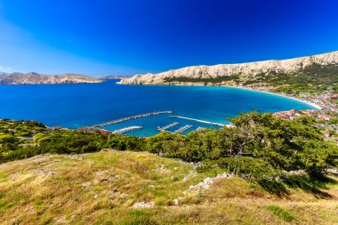 Hırvatistan 'ın Krk Adası' ndaki kayalık dağlarla çevrili turkuaz Adriyatik Denizi, marina, kumlu plaj ve kıyı kasabasının açık mavi gökyüzü altında Baska Körfezi manzaralı manzarası.