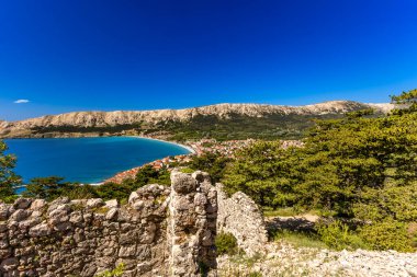 Hırvatistan 'ın Krk Adası' ndaki kayalık dağlarla çevrili turkuaz Adriyatik Denizi, marina, kumlu plaj ve kıyı kasabasının açık mavi gökyüzü altında Baska Körfezi manzaralı manzarası.