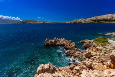 Hırvatistan 'ın Krk Adası' ndaki kayalık dağlarla çevrili turkuaz Adriyatik Denizi, marina, kumlu plaj ve kıyı kasabasının açık mavi gökyüzü altında Baska Körfezi manzaralı manzarası.