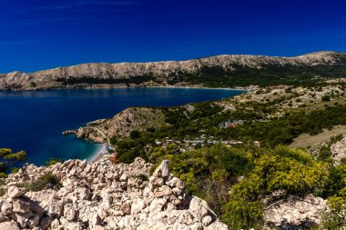 Hırvatistan 'ın Krk Adası' ndaki kayalık dağlarla çevrili turkuaz Adriyatik Denizi, marina, kumlu plaj ve kıyı kasabasının açık mavi gökyüzü altında Baska Körfezi manzaralı manzarası.