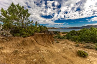 Engebeli kayalık zemin, yeşil çalılar, çam ağaçları ve dağınık bulutlu dramatik bir gökyüzü altında vahşi bitki örtüsü barındıran Akdeniz manzarasının güzel panoramik manzarası. Sahne sakin mavi bir denize bakıyor. Uzakta dağlar var.
