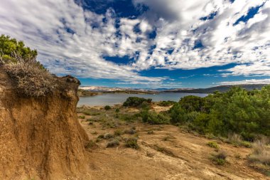 Engebeli kayalık zemin, yeşil çalılar, çam ağaçları ve dağınık bulutlu dramatik bir gökyüzü altında vahşi bitki örtüsü barındıran Akdeniz manzarasının güzel panoramik manzarası. Sahne sakin mavi bir denize bakıyor. Uzakta dağlar var.