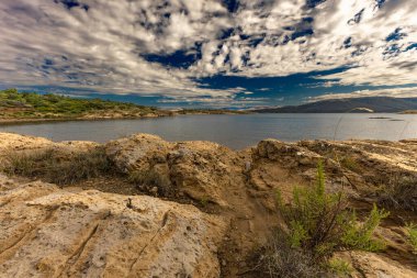 Engebeli kayalık zemin, yeşil çalılar, çam ağaçları ve dağınık bulutlu dramatik bir gökyüzü altında vahşi bitki örtüsü barındıran Akdeniz manzarasının güzel panoramik manzarası. Sahne sakin mavi bir denize bakıyor. Uzakta dağlar var.