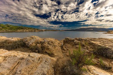 Engebeli kayalık zemin, yeşil çalılar, çam ağaçları ve dağınık bulutlu dramatik bir gökyüzü altında vahşi bitki örtüsü barındıran Akdeniz manzarasının güzel panoramik manzarası. Sahne sakin mavi bir denize bakıyor. Uzakta dağlar var.