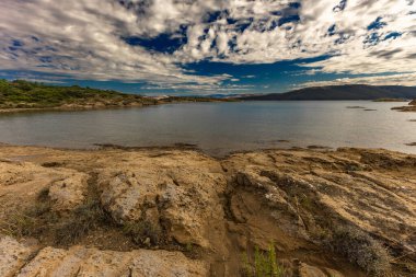 Engebeli kayalık zemin, yeşil çalılar, çam ağaçları ve dağınık bulutlu dramatik bir gökyüzü altında vahşi bitki örtüsü barındıran Akdeniz manzarasının güzel panoramik manzarası. Sahne sakin mavi bir denize bakıyor. Uzakta dağlar var.