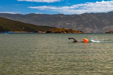 Active swimmer training in open water with a bright orange safety buoy, creating splashes on the surface of the sea. The scene is set against a stunning natural backdrop of mountains and rocky coastline under a blue sky with scattered clouds. Perfect