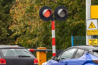 Kırmızı ve gri dizel bir lokomotif geçerken arabalar kapalı bir demiryolu geçidinde durur. Güvenlik bariyerleri ve uyarı ışıkları bulutlu bir günde yaklaşan trene işaret ediyor..