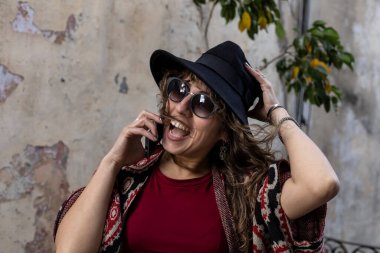 Young Latin American woman with curly hair, hat, sunglasses, positive attitude talks on cell phone
