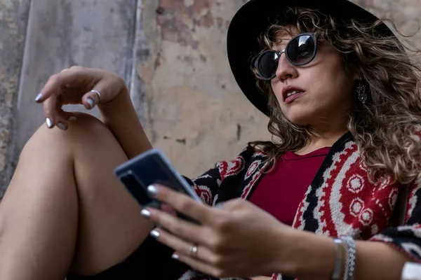 Young latina woman with curly hair, hat, sunglasses, relaxed attitude with cell phone in her hand