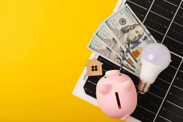 Flat lay composition with solar panel, LED lamp, piggy bank, house model and money on yellow background. Money saving and clean energy concept. Ecology and sustainable development concept. Place for text