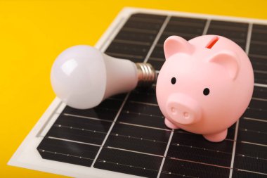 Güneş paneli, öncü lamba ve sarı arka planda domuz kumbarası olan düz bir kompozisyon. Tasarruf ve temiz enerji kavramı. Ekoloji ve sürdürülebilir kalkınma kavramı.