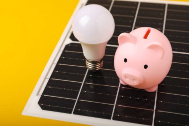 Güneş paneli, öncü lamba ve sarı arka planda domuz kumbarası olan düz bir kompozisyon. Tasarruf ve temiz enerji kavramı. Ekoloji ve sürdürülebilir kalkınma kavramı.