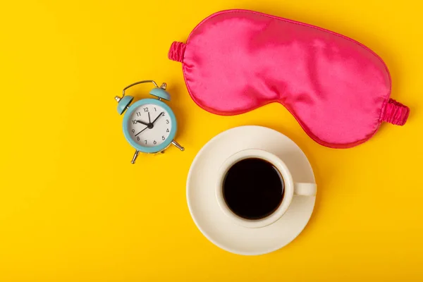 Sleeping mask, alarm clock and a cup of coffee on a yellow background. The concept of cheerful morning, GOOD MORNING, good night, insomnia and relaxation. View from above. flat lay