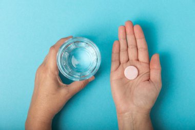 Kadının elleri mavi zeminde vitamin hapı ve bir bardak su tutuyor. Baş ağrısı ve soğuk için ilaç almaya hazır. Ağrı kesiciler, besin takviyeleri, bağışıklık hapı..
