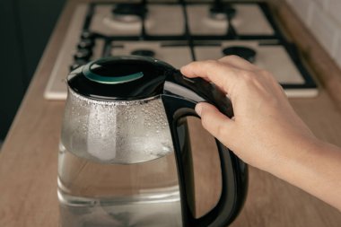 Mutfaktaki bir masada elektrikli su ısıtıcısının elektrik düğmesine basan bir kadının yakın çekimi. Su kaynatmak, çay ve kahve yapmak için kettle. Sıcak içecekler için ev aletleri. Evde enerji tasarrufu..