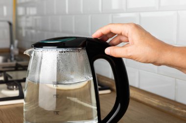 Mutfaktaki bir masada elektrikli su ısıtıcısının elektrik düğmesine basan bir kadının yakın çekimi. Su kaynatmak, çay ve kahve yapmak için kettle. Sıcak içecekler için ev aletleri. Evde enerji tasarrufu..