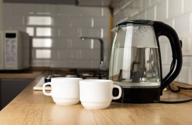 Mutfaktaki masada çay içmek için kaynar su ve fincanlarla şeffaf elektrikli su ısıtıcısı..