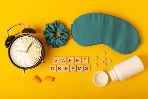 Sleep mask, alarm clock, earplugs, melatonin pills for insomnia and text SWEET DREAMS on a yellow background. The concept of rest, sleep quality, good night, insomnia and relaxation.Flat lay, mockup.