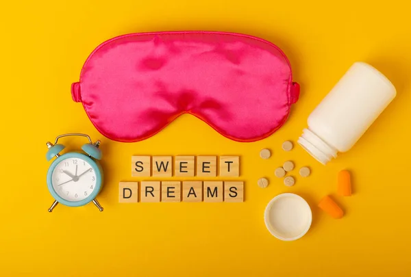 Sleep mask, alarm clock, earplugs, melatonin pills for insomnia and text SWEET DREAMS on a yellow background. The concept of rest, sleep quality, good night, insomnia and relaxation.Flat lay, mockup.