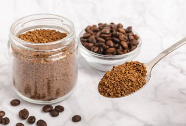 Instant coffee beans in a glass jar on a white marble background. Hot drink ingredients. Instant coffee on the table. Espresso. Place for text. Place to copy. Banner.