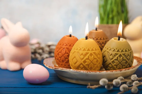 Easter candle eggs in a plate, Easter bunny and willow branches on a blue table and a blurred light background. Easter holiday concept.Spring willow bouquet.Decor for home and festive steel.Copy space. Place for text.