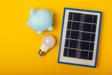 Güneş paneli, öncü lamba ve sarı arka planda domuz kumbarası olan düz bir kompozisyon. Tasarruf ve temiz enerji kavramı. Ekoloji ve sürdürülebilir kalkınma kavramı.