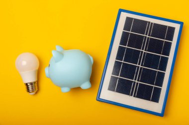 Güneş paneli, öncü lamba ve sarı arka planda domuz kumbarası olan düz bir kompozisyon. Tasarruf ve temiz enerji kavramı. Ekoloji ve sürdürülebilir kalkınma kavramı.