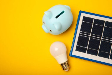 Güneş paneli, öncü lamba ve sarı arka planda domuz kumbarası olan düz bir kompozisyon. Tasarruf ve temiz enerji kavramı. Ekoloji ve sürdürülebilir kalkınma kavramı.