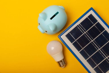 Güneş paneli, öncü lamba ve sarı arka planda domuz kumbarası olan düz bir kompozisyon. Tasarruf ve temiz enerji kavramı. Ekoloji ve sürdürülebilir kalkınma kavramı.